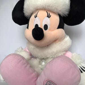 May include: Pink Minnie Mouse plush toy wearing a white fur trimmed hat and pink snowsuit with a silver zipper. The plush toy has black embroidered eyes and a black nose. The toy has a Disney Store Exclusive tag on the foot.