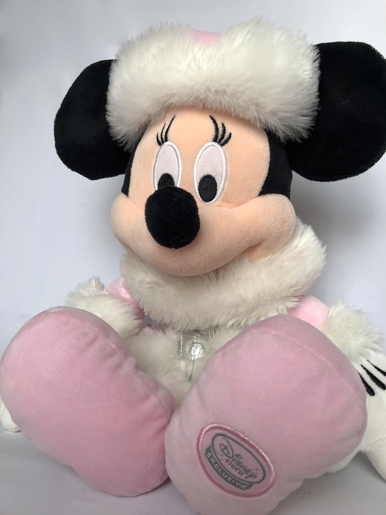 May include: Pink Minnie Mouse plush toy wearing a white fur trimmed hat and pink snowsuit with a silver zipper. The plush toy has black embroidered eyes and a black nose. The toy has a Disney Store Exclusive tag on the foot.
