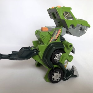 May include: Green and grey toy dinosaur that transforms into a construction vehicle. The toy has wheels and a claw. The toy has the words "Switch & Go Dino" printed on the side.
