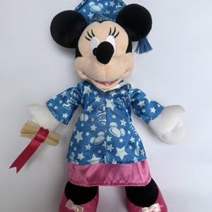 May include: A Minnie Mouse plush toy wearing a blue graduation gown with white stars and a matching cap. The plushie holds a diploma with a red ribbon and wears pink shoes with bows. The graduation gown has the text "2024".