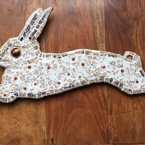 Peut inclure: Sculpture de lapin en céramique blanche avec des accents marrons et bleus. Le lapin saute en avant, les oreilles dressées.