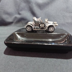 May include: A silver toy car sits on a black ceramic tray. The car has a detailed design and appears to be an antique.