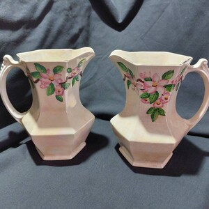 Peut inclure: Deux pichets en céramique blanche avec des motifs floraux roses et verts. Les pichets ont une forme hexagonale et une poignée d'un côté.