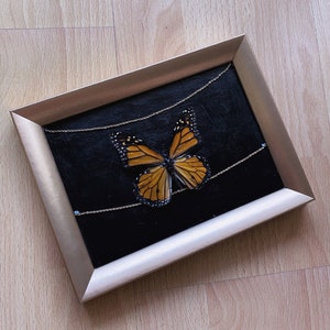 Puede incluir: Una obra de arte enmarcada que presenta una mariposa con alas naranja y negras sobre un fondo negro. La mariposa está rodeada de delicadas cadenas dorées.