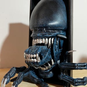 May include: A dark blue Alien sculpture with a black frame. The sculpture features a detailed head with sharp teeth and a textured surface. The creature's limbs extend outward, showcasing sharp claws. The overall design is a representation of the Alien creature.