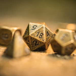 Puede incluir: Primer plano de un conjunto de dados de metal dorado, incluyendo un dado de 20 caras con los números 9, 18 y 4 visibles. Los dados tienen una superficie texturizada y son perfectos para juegos de mesa.