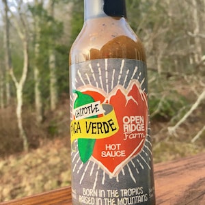 May include: A glass bottle of Pica Verde Chipotle hot sauce with a gray and red label. The label features a green pepper, a red heart, and the text "Open Ridge Farm". The bottle is sitting on a wooden surface.