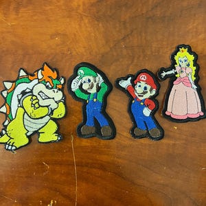 Peut inclure: Quatre patchs brodés représentant des personnages de la série de jeux vidéo Super Mario Bros. Les patchs sont sur une surface en bois brun. Les personnages sont Bowser, Luigi, Mario et la princesse Peach.