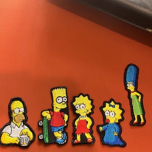 Può includere: Sei toppe ricamate da stirare con la famiglia Simpson. Le toppe sono gialle, blu, rosse e verdi. I personaggi includono Homer, Bart, Marge, Lisa e Maggie.