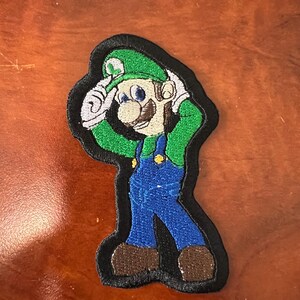 May include: Green and blue embroidered patch of a cartoon character with a green hat and mustache. The character is wearing blue overalls and has a black outline.
