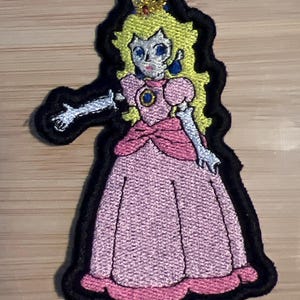 Könnte beinhalten: Ein gestickter Bügel-Patch mit einem Cartoon-Bild von Prinzessin Peach aus der Super Mario Bros. Videospielserie. Sie trägt ein rosa Kleid mit einem blauen Edelstein und einer gelben Krone.