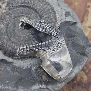 Peut inclure: Bague en argent en forme de tête d'alligator, avec des écailles détaillées et une gueule ouverte. La bague est posée sur un fond de pierre grise texturée.