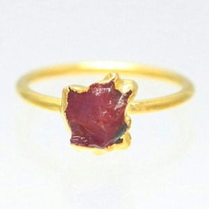 May include: A gold ring with a raw ruby gemstone. The ruby is a deep red color and has a rough, natural shape.