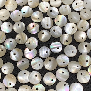 May include: A collection of round, iridescent mother-of-pearl shell beads. Each bead has a small hole in the center, and the shells display a range of colors, including white, pink, and green. The beads are scattered on a dark surface.
