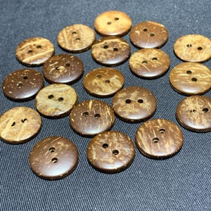 May include: A selection of round coconut shell buttons in natural tones. The buttons, in shades of brown and tan, each have two or three holes for sewing. They are arranged on a dark grey fabric surface.