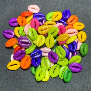 May include: A selection of vibrant cowrie shell beads in a range of colours such as orange, purple, green, yellow, pink, and blue. These glossy, oval-shaped beads have a slit opening, ideal for jewellery making or craft projects.