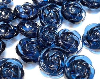 May include: A collection of dark blue glass rose-shaped buttons. The buttons have a detailed, swirling petal design and a glossy finish. The buttons are scattered across a white surface, creating a visually appealing arrangement.