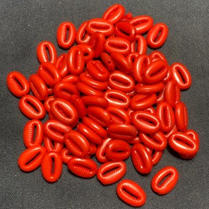 Puede incluir: Un montón de conchas de cauri de color rojo brillante, de forma ovalada con una abertura en forma de hendidura, se encuentran sobre una superficie gris oscuro. Las conchas son lisas y brillantes, con un color uniforme. Estas conchas se utilizan a menudo en joyería y artesanía.