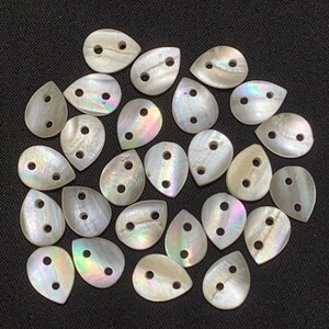 May include: A collection of iridescent, teardrop-shaped shell buttons with two holes each. The buttons have a pearlescent sheen, reflecting colors of pink, green, and blue. They are scattered on a dark background.