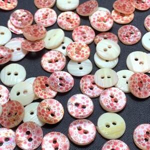 May include: A collection of round buttons in various shades of white and pink. Some buttons feature a floral pattern in pink and red, while others are solid white. Each button has two or four holes for sewing.
