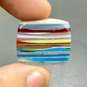 May include: A square, colorful glass bead with horizontal stripes in various colors, including red, blue, yellow, and white. The bead is held between two fingers, showcasing its polished surface and layered design. The bead is approximately 1 inch square.
