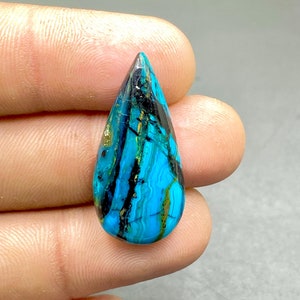 Könnte beinhalten: Ein tropfenförmiger Cabochon-Edelstein mit einem blauen, grünen und schwarzen Muster. Der Stein hat eine glatte, polierte Oberfläche.