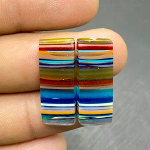 May include: Two rectangular, colorful glass cabochons with horizontal stripes in shades of yellow, orange, red, blue, and green. The cabochons are held between fingers, showcasing their vibrant, layered design. These are suitable for jewelry making.