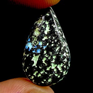 May include: A teardrop-shaped gemstone with a black base, speckled with green, white, and blue inclusions. The polished surface reflects light, highlighting the unique patterns and colours. The gemstone is held between two fingers against a black background.