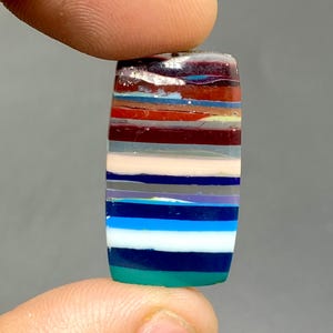 May include: A multi-colored, layered, rectangular cabochon gemstone with a variety of colors including red, blue, green, white, and gray.