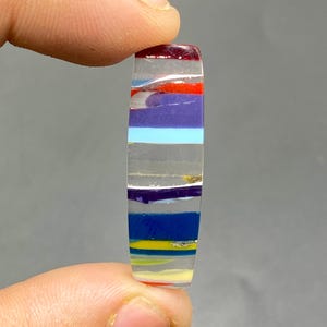 May include: A multi-colored glass cabochon with a striped pattern. The cabochon is clear with red, orange, yellow, blue, purple, and white stripes.