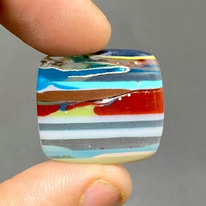May include: A square, multi-colored glass cabochon with layers of blue, turquoise, brown, red, white, and green glass. The glass has a layered, striated appearance.