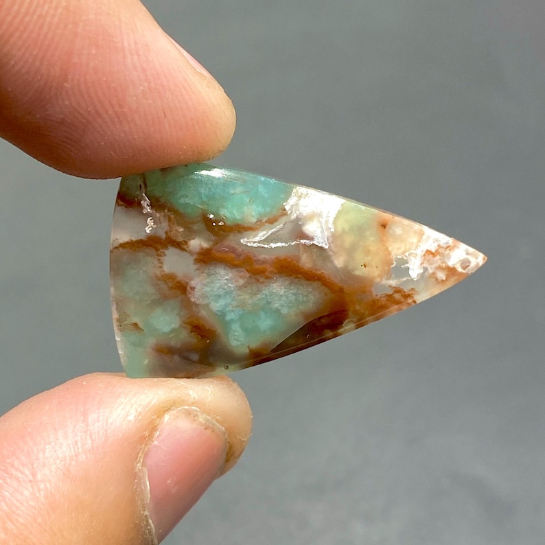 May include: A triangular shaped cabochon of green, brown, and white agate. The stone has a mottled pattern and is translucent.