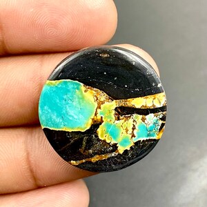Puede incluir: Una piedra preciosa redonda con un contraste de colores llamativo. La piedra presenta una gran área negra, con inclusiones turquesas y amarillo-dorado vibrantes. La superficie pulida refleja la luz, resaltando los patrones y texturas naturales.