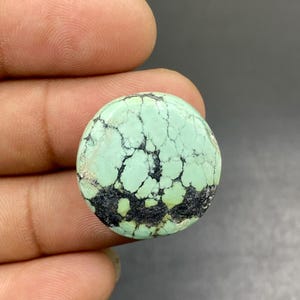 Pode incluir: Um cabochão de pedra preciosa turquesa de forma redonda, apresentando uma superfície verde-menta pálida com veios de matriz pretos intrincados e uma inclusão proeminente de cor carvão escuro ao longo da borda inferior, com acabamento liso e brilhante.