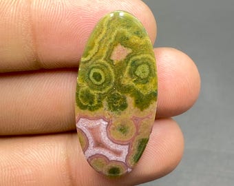 May include: Oval-shaped gemstone with a unique pattern. The stone features a blend of green, yellow, and pink hues, with circular and swirling patterns. The polished surface reflects light, highlighting the natural variations in color and texture. A beautiful, natural stone.