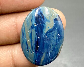 May include: Oval-shaped gemstone with a swirling pattern of deep blue, light blue, and gray hues. Tiny white speckles are scattered across the surface, resembling stars. The polished stone has a glossy finish.