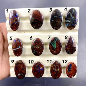 May include: A collection of twelve polished gemstones, each with unique patterns and colors. The stones are primarily oval-shaped, with a few teardrop forms. Colors include deep reds, browns, blues, and greens, creating a marbled effect. The gemstones are displayed on a white, ridged surface.