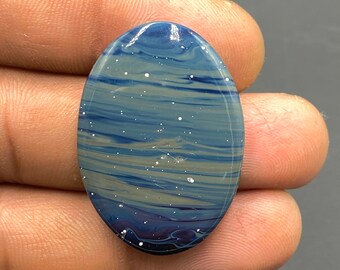 May include: An oval-shaped gemstone with a swirling pattern of blue, gray, and white. The polished surface has a glossy finish, with small white flecks scattered across the stone. The gemstone is held between fingers.