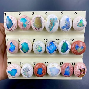 Op de afbeelding: Een verzameling van achttien ovale opaal edelstenen, elk geplaatst op een crèmekleurige display. De opalen tonen een verscheidenheid aan kleuren, waaronder blauw, groen, roze en wit. De edelstenen zijn genummerd van 1 tot 18.