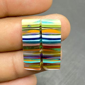 May include: Two rectangular, colorful glass cabochons with a striped pattern. The cabochons feature layers of various colors, including yellow, orange, blue, and purple. The cabochons are held between fingers against a gray background.