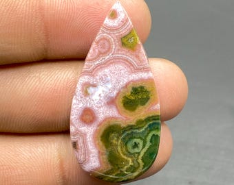 May include: A teardrop-shaped cabochon of pink, green, and white ocean jasper. The stone has a swirling pattern of concentric circles and irregular shapes.