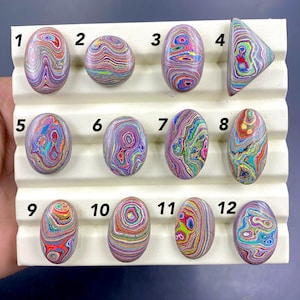 May include: Twelve polished gemstones in diverse shapes, including oval and triangular. The stones exhibit a vibrant array of colours, such as red, blue, green, and purple, with unique swirling patterns. They are arranged on a white display.