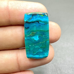 Op de afbeelding: Een rechthoekige edelsteen met een gepolijst oppervlak. De steen heeft een mix van turquoise, blauwgroene en diepblauwe tinten, met zwarte strepen en hints van groen. De edelsteen wordt tussen vingers gehouden.