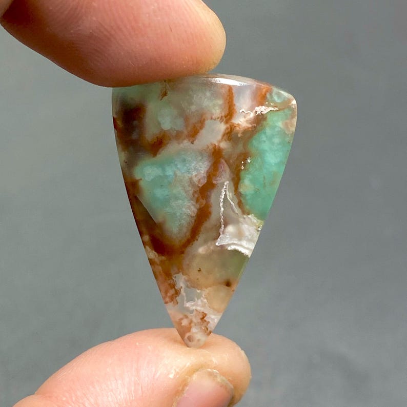 May include: A green, brown, and white gemstone cabochon with a triangular shape. The gemstone has a unique pattern of swirling colors and textures.