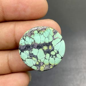 Può includere: Un cabochon di pietra turchese rotondo e levigato, caratterizzato da un vibrante colore verde menta con un prominente motivo a matrice scura e reticolata che attraversa orizzontalmente il centro e si diffonde sulla superficie liscia.