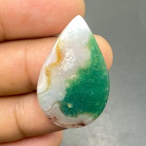 May include: A polished, teardrop-shaped gemstone. The stone displays a blend of colours: white, green, and touches of yellow and brown. The gemstone is held between fingers, highlighting its form and size.