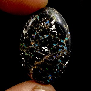 May include: An oval-shaped black opal gemstone featuring a dark base colour with scattered, vibrant flecks of blue, green, and white iridescent inclusions throughout the surface, polished to a smooth, reflective finish with a high-gloss sheen.