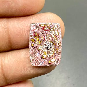 May include: A rectangular gemstone with a unique pattern of red, yellow, white, and brown swirls and spots. The stone has a polished surface and is held between fingers, showcasing its intricate design and color variations. The gemstone is likely for jewelry making.