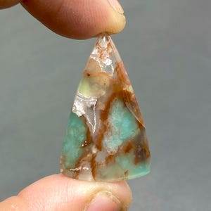 May include: A triangular shaped pendant made of green, brown, and white agate. The pendant has a unique pattern of swirling colors and textures.