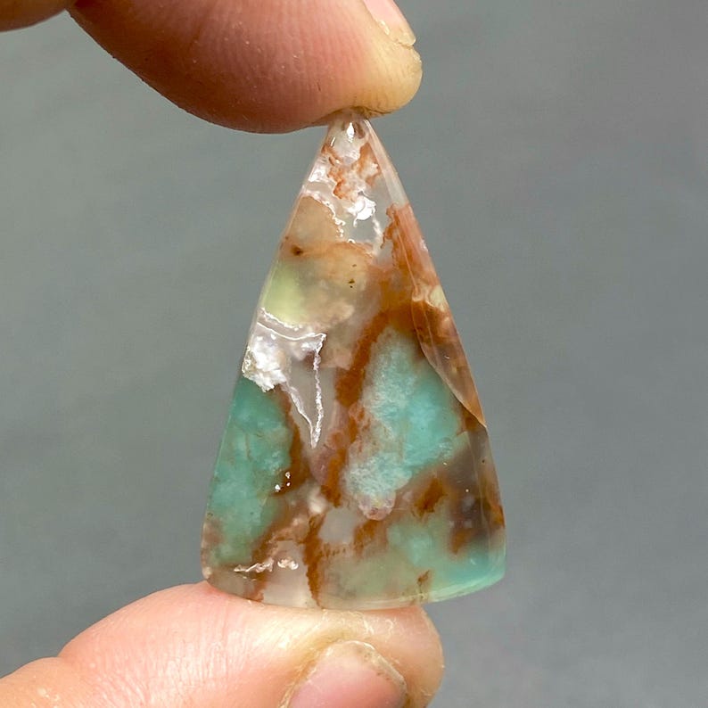 May include: A triangular shaped pendant made of green, brown, and white agate. The pendant has a unique pattern of swirling colors and textures.